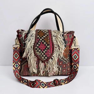 May include: A woven handbag with a colorful geometric pattern and cream-colored fringe. The bag has black handles and a detachable shoulder strap with a repeating diamond design. The bag is approximately 12 inches wide.