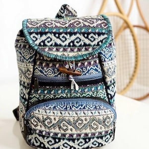 May include: A small backpack with a patterned design in shades of blue, purple, and cream. The bag features a flap closure with a wooden toggle, a front zippered pocket, and a teal trim. The backpack is approximately 12 inches tall.