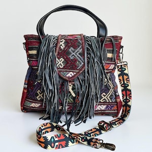 May include: A colorful handbag with a patterned textile body, black leather handles, and a long, fringed flap. The bag includes a detachable, patterned shoulder strap. The textile design features geometric shapes in red, purple, and orange.