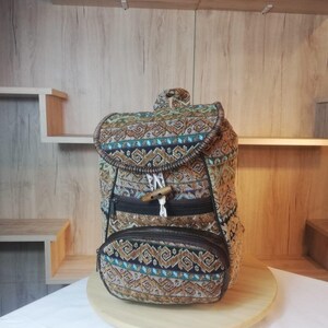 Könnte beinhalten: Ein Rucksack aus gewebtem Stoff mit geometrischem Muster in Braun-, Beige-, Blau- und Schwarztönen. Die Tasche verfügt über eine obere Klappe mit Holzknebelverschluss, eine Reißverschlusstasche auf der Vorderseite und dunkelbraune Kanten.