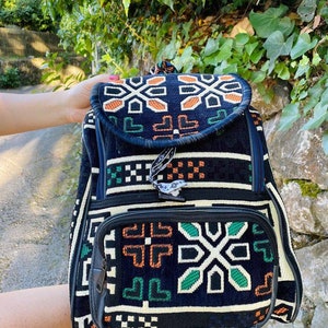 May include: Black backpack with a geometric pattern in green, orange, and white. The backpack has a flap with a zipper closure and a smaller pocket on the front.