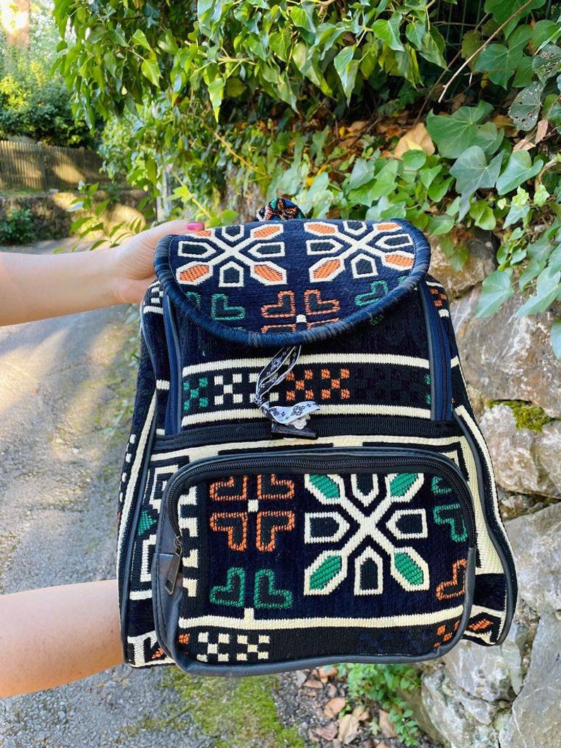 May include: Black backpack with a geometric pattern in green, orange, and white. The backpack has a flap with a zipper closure and a smaller pocket on the front.