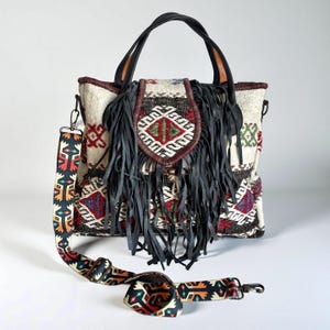 May include: A cream-colored tote bag with a patterned design and a black leather fringe. The bag has a patterned shoulder strap and black handles. The bag's design includes red, green, and blue geometric shapes.