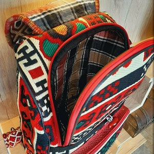 May include: A red, black and white patterned backpack with a red zipper and a black leather handle. The backpack is made of woven fabric and has a traditional design.
