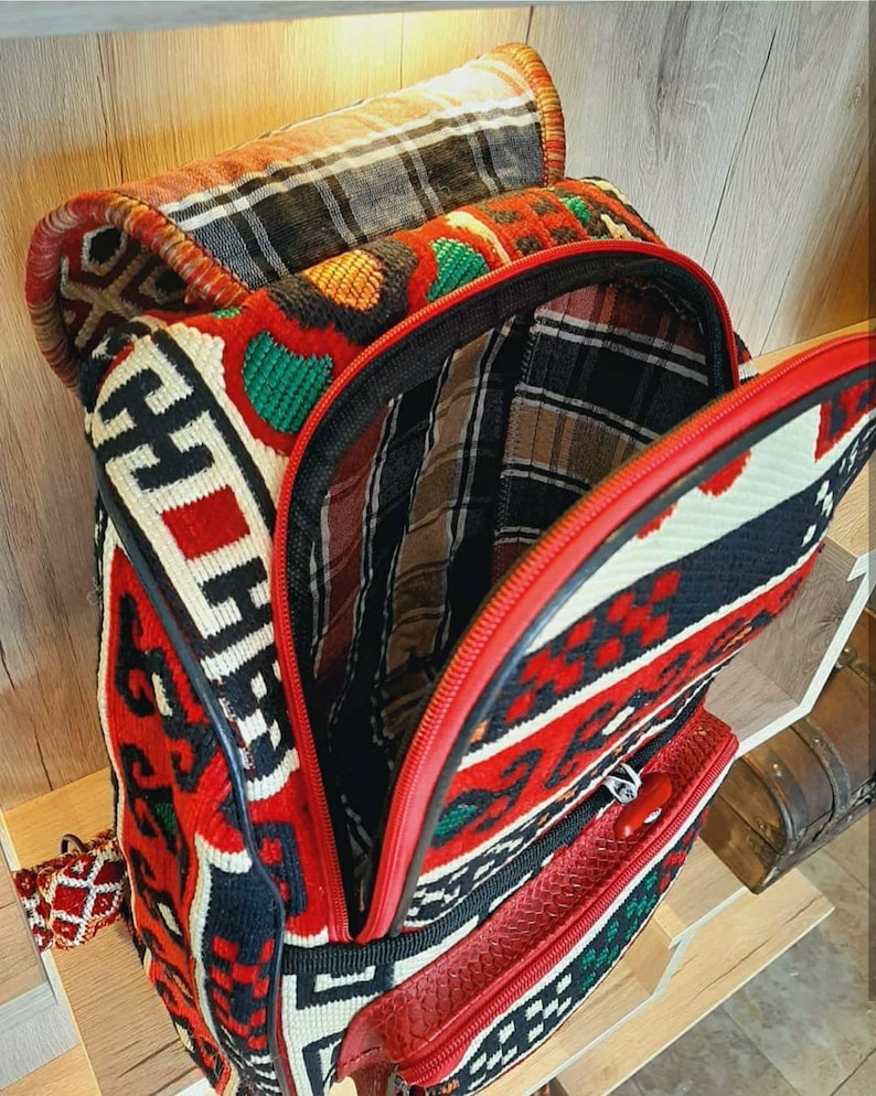 May include: A red, black and white patterned backpack with a red zipper and a black leather handle. The backpack is made of woven fabric and has a traditional design.