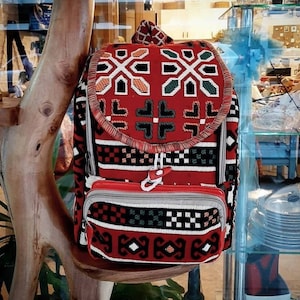 May include: A backpack featuring a red, black, and white woven geometric pattern. The bag includes a top flap with a drawstring closure, a front zippered pocket, and grey trim. The fabric displays intricate repeating shapes and lines.