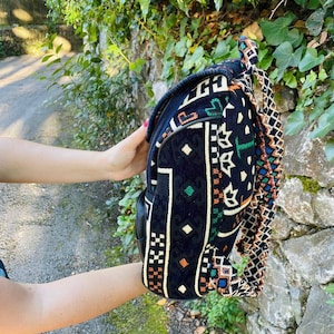May include: A black backpack with a colorful geometric pattern and a long, braided strap. The backpack is made of a woven fabric and has a zipper closure.