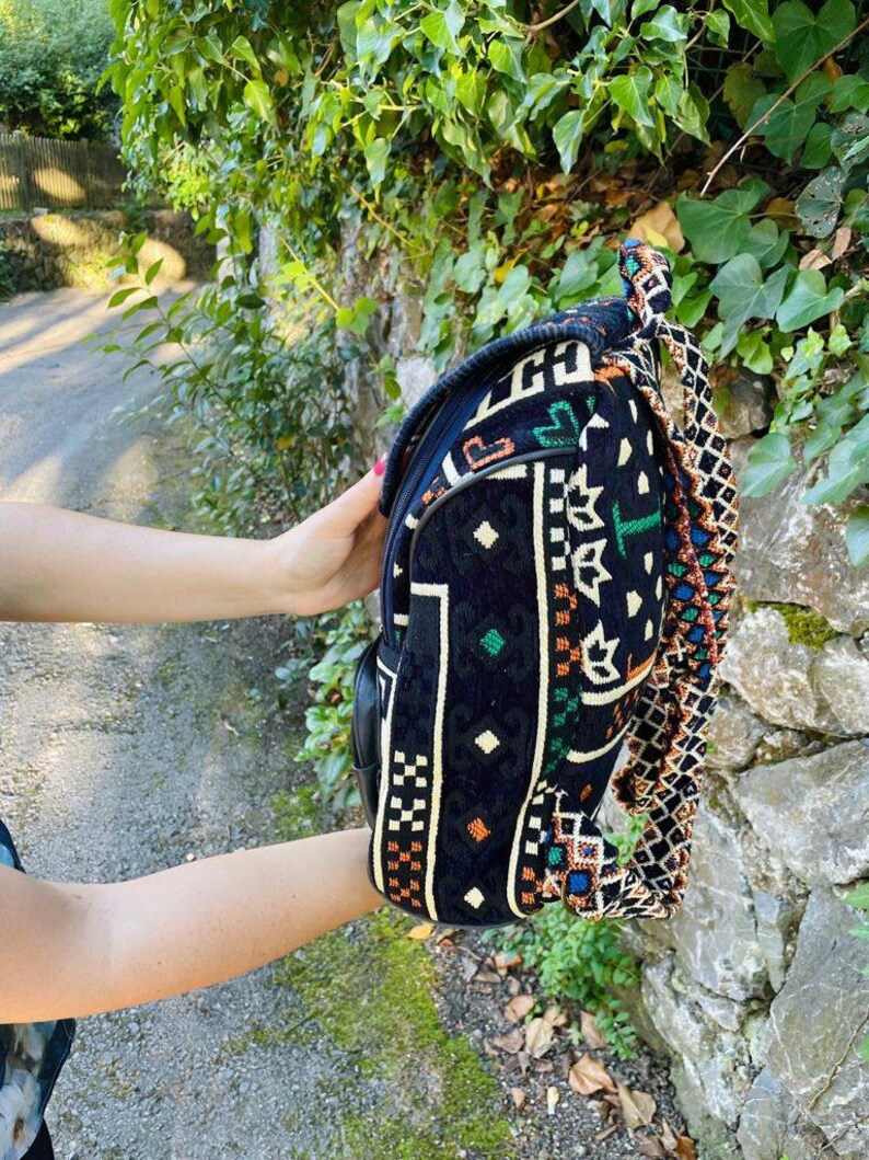 May include: A black backpack with a colorful geometric pattern and a long, braided strap. The backpack is made of a woven fabric and has a zipper closure.