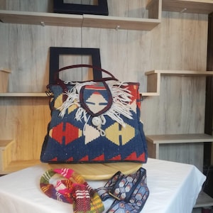 May include: A rectangular tote bag featuring a blue, red, and yellow geometric woven pattern with a central flap adorned with white fringe and a circular button closure. Two detachable straps are included: one multicolored and one blue patterned.