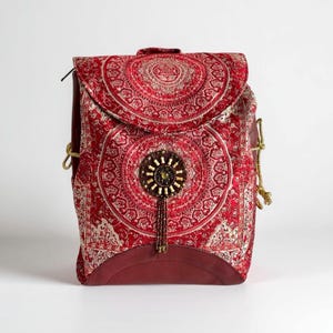 Puede incluir: Mochila roja con un diseño intrincado y estampado. La solapa presenta un motivo circular grande, y la bolsa está adornada con un adorno de cuentas. La mochila tiene una base y un ribete burdeos, y correas de cuerda doradas.