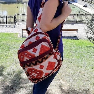 Könnte beinhalten: Ein Rucksack aus gewebtem Stoff mit geometrischem Muster in Orange-, Creme- und Dunkelbrauntönen. Die Tasche verfügt über ein vorderes Reißverschlussfach, eine Hauptöffnung mit Reißverschluss und breite Schultergurte aus demselben gemusterten Stoff.