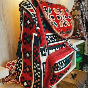 May include: A red, black and white patterned backpack with a brown leather strap. The backpack has a zipped pocket on the front and a large main compartment.