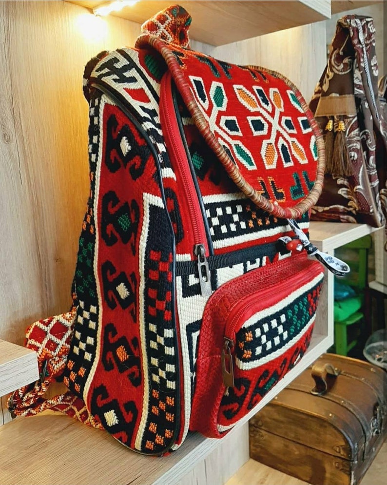 May include: A red, black and white patterned backpack with a brown leather strap. The backpack has a zipped pocket on the front and a large main compartment.