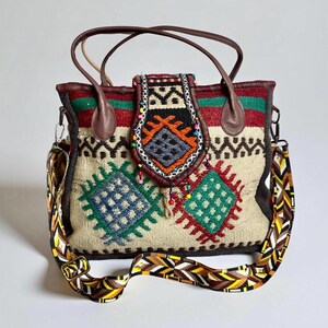 May include: A beige tote bag with a colorful woven design. The bag features brown leather handles and a detachable patterned shoulder strap. The front of the bag has embroidered geometric shapes in blue, green, and red. The top flap has a decorative beaded trim.