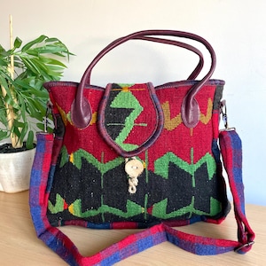 Könnte beinhalten: Gewebte Handtasche mit rotem, schwarzem und grünem geometrischem Muster. Die Tasche hat einen bordeauxfarbenen Ledergriff und Akzente, einen Holzknopfverschluss und einen karierten Schulterriemen. Die Tasche steht auf einer Holzoberfläche.