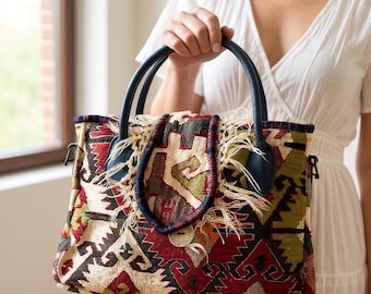 May include: A rectangular handbag crafted from woven fabric featuring a multi-colored geometric pattern in shades of red, green, cream, and black. The bag includes dark blue leather handles, frayed fabric edges, and a single wooden button closure.