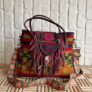 May include: A red, green, and blue woven tapestry tote bag with a burgundy leather fringe and a brown leather strap. The bag has a woven pattern and a button closure.