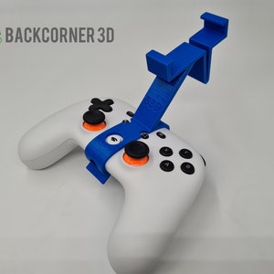 May include: A white games controller with black buttons and orange accents, held by a blue 3D-printed phone holder. The holder is attached to the controller and features the text "B3D". The logo "BACKCORNER 3D" is in the upper left corner.