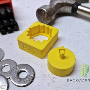 Puede incluir: Una herramienta de estampado de metal impresa en 3D de color amarillo con las letras "B3D" en el lateral. La herramienta tiene una abertura circular y una pieza separada impresa en 3D de color amarillo con un peque&ntilde;o poste en el centro. Hay cuatro arandelas plateadas en la imagen.