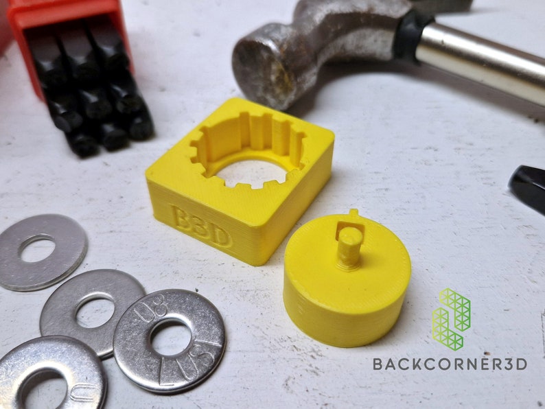 Puede incluir: Una herramienta de estampado de metal impresa en 3D de color amarillo con las letras "B3D" en el lateral. La herramienta tiene una abertura circular y una pieza separada impresa en 3D de color amarillo con un peque&ntilde;o poste en el centro. Hay cuatro arandelas plateadas en la imagen.