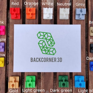 Puede incluir: Una colecci&oacute;n de 14 peque&ntilde;as piezas de rompecabezas de pl&aacute;stico en varios colores, incluyendo rojo, naranja, blanco, neutro, gris, rosa, morado, amarillo, bronce, negro, azul oscuro, dorado, verde claro, verde oscuro y azul claro. Las piezas est&aacute;n dispuestas en dos filas sobre una superficie de madera. El texto "BACKCORNER 3D" est&aacute; impreso en una tarjeta blanca en el centro de la imagen.