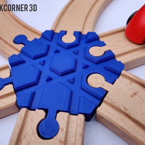 Può includere: Un connettore blu a forma di fiocco di neve per binari del treno, stampato in 3D. Il connettore è posizionato tra i binari del treno in legno, con un'auto giocattolo rossa visibile. Il logo "BACKCORNER 3D" è in alto a sinistra.