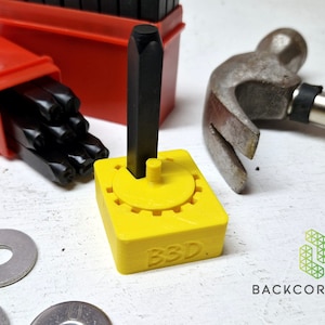 Puede incluir: Una herramienta amarilla impresa en 3D para sujetar un punz&oacute;n de letras de metal. La herramienta tiene un mecanismo de engranaje giratorio para cambiar el punz&oacute;n de letras. Un punz&oacute;n de letras de metal negro est&aacute; en la herramienta. Un martillo y una caja de punzones de letras est&aacute;n en el fondo.