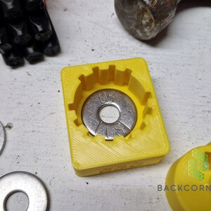 Puede incluir: Un molde amarillo impreso en 3D para una arandela de metal. El molde tiene un borde elevado alrededor del borde y una depresi&oacute;n circular en el centro. Una arandela plateada con el texto "US" y "0.5" est&aacute; dentro del molde. Dos arandelas plateadas m&aacute;s son visibles en la superficie blanca.