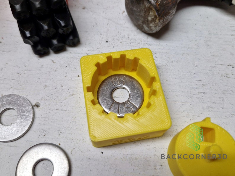 Puede incluir: Un molde amarillo impreso en 3D para una arandela de metal. El molde tiene un borde elevado alrededor del borde y una depresi&oacute;n circular en el centro. Una arandela plateada con el texto "US" y "0.5" est&aacute; dentro del molde. Dos arandelas plateadas m&aacute;s son visibles en la superficie blanca.