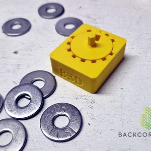 Puede incluir: Un dispositivo amarillo impreso en 3D con un mecanismo de engranaje rotatorio y el texto "B3D" en el lateral. El dispositivo est&aacute; rodeado de varias arandelas de metal plateado.