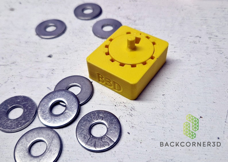 Puede incluir: Un dispositivo amarillo impreso en 3D con un mecanismo de engranaje rotatorio y el texto "B3D" en el lateral. El dispositivo est&aacute; rodeado de varias arandelas de metal plateado.