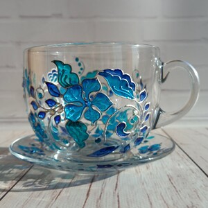 May include: A clear glass teacup and saucer set. The cup and saucer are decorated with a hand-painted floral design in shades of turquoise and blue. The floral design includes flowers, leaves, and vines. The cup has a handle.