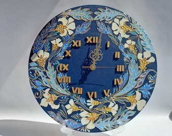 May include: A round, navy blue wall clock with gold Roman numerals and hands. The clock face is decorated with a floral design in white, gold, and shades of blue. The clock is displayed on a clear stand.