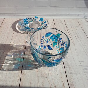 May include: A clear glass teacup and saucer set. The cup and saucer are decorated with hand-painted blue floral designs. The cup has a handle and sits next to the saucer on a light wood surface. The set is illuminated by sunlight.