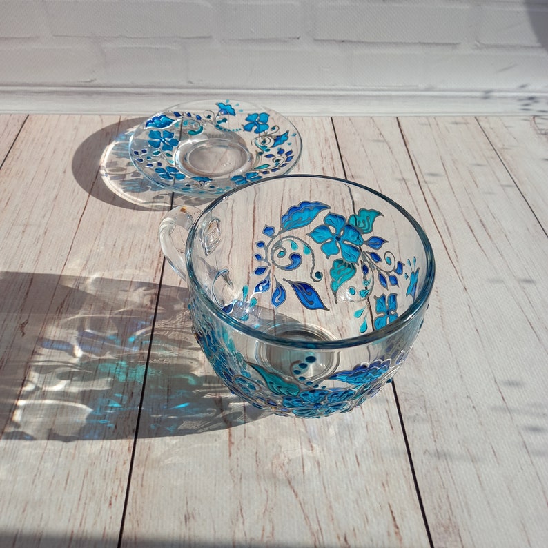May include: A clear glass teacup and saucer set. The cup and saucer are decorated with hand-painted blue floral designs. The cup has a handle and sits next to the saucer on a light wood surface. The set is illuminated by sunlight.