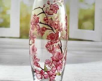May include: A clear glass vase with an oval silhouette, decorated with a hand-painted design of pink cherry blossoms on dark brown branches. The floral pattern features translucent pink petals with white highlights and gold-toned outlines.