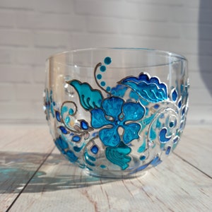 May include: A clear glass bowl with hand-painted floral designs in shades of turquoise and blue. The flowers and leaves are outlined in silver, creating a decorative and artistic piece. The bowl is ideal for serving or display.