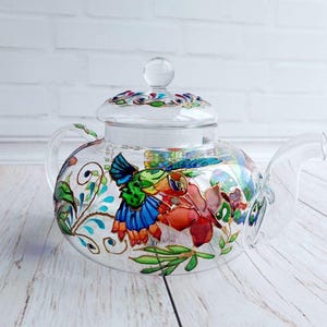 Puede incluir: Una tetera de vidrio transparente con tapa y asa. La tetera est&aacute; decorada con coloridos dise&ntilde;os florales y de aves en tonos azules, verdes, rojos y naranjas. La tetera se encuentra sobre una superficie de madera clara.