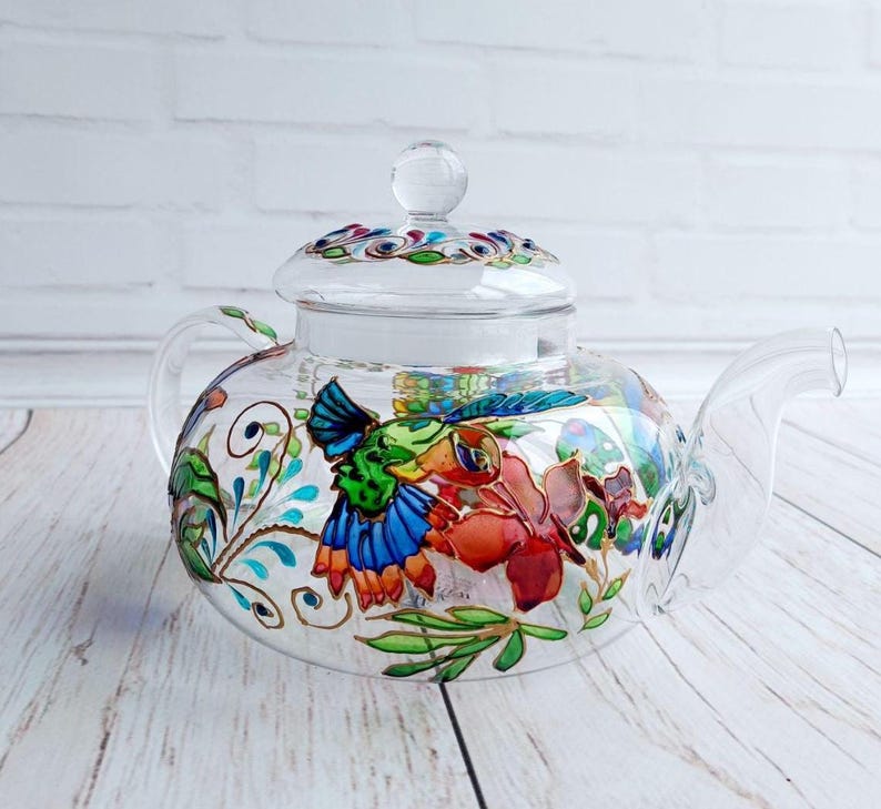 Puede incluir: Una tetera de vidrio transparente con tapa y asa. La tetera est&aacute; decorada con coloridos dise&ntilde;os florales y de aves en tonos azules, verdes, rojos y naranjas. La tetera se encuentra sobre una superficie de madera clara.