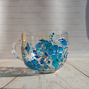 May include: Clear glass teacup with a large handle, hand-painted with a floral design in shades of turquoise and blue. The flowers and leaves are outlined in silver, creating a decorative and elegant look. The cup sits on a light wood surface.