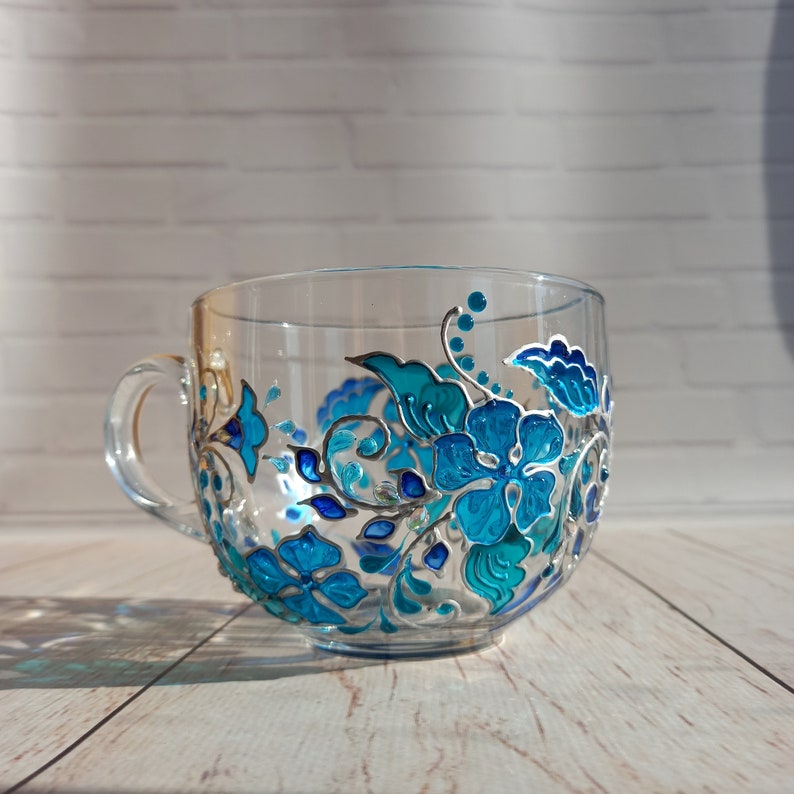 May include: Clear glass teacup with a large handle, hand-painted with a floral design in shades of turquoise and blue. The flowers and leaves are outlined in silver, creating a decorative and elegant look. The cup sits on a light wood surface.