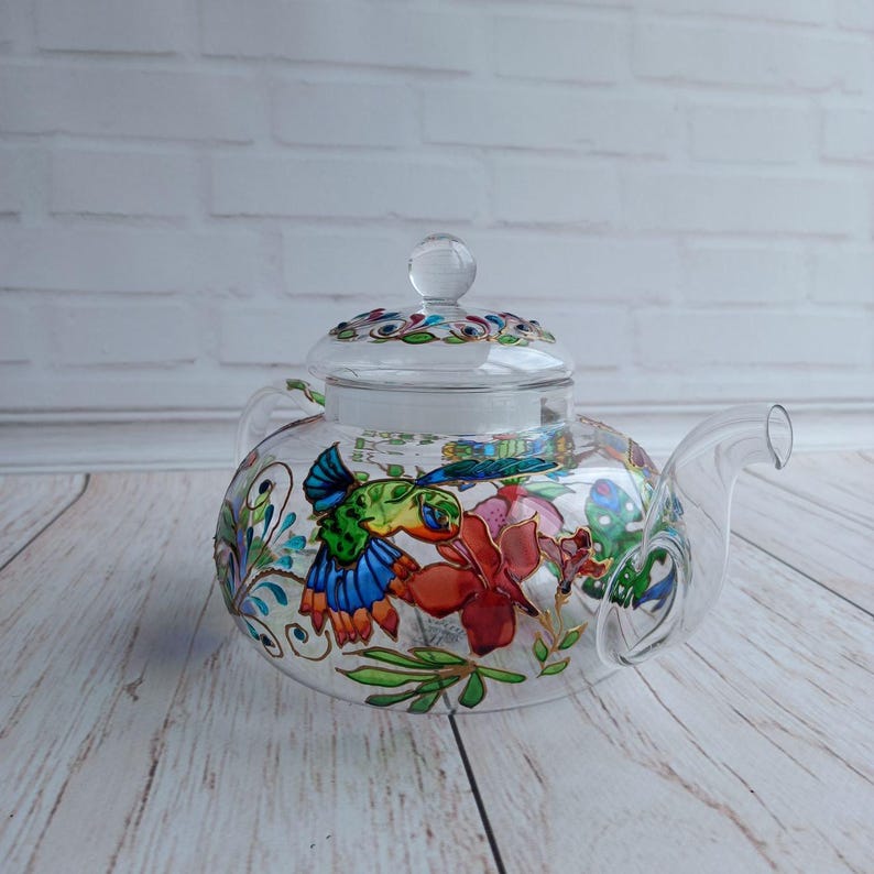 Puede incluir: Una tetera de vidrio transparente con tapa, asa y boquilla. La tetera est&aacute; decorada con coloridos dise&ntilde;os florales y de p&aacute;jaros. La tapa tiene un pomo de vidrio transparente. La tetera est&aacute; sobre una superficie de madera clara.