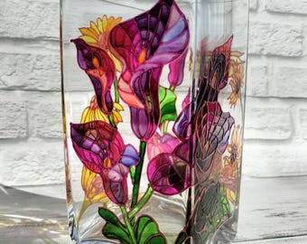 May include: A clear, rectangular glass vase with hand-painted floral designs. The vase features vibrant purple, pink, and yellow calla lilies and other flowers. Green leaves and stems add contrast. The vase is a decorative home accent.