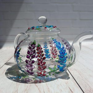 May include: A clear glass teapot with a painted design of purple, pink, blue, and green lupine flowers.