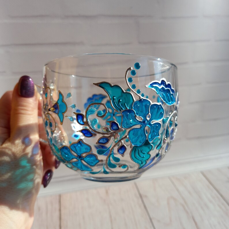 May include: A clear glass teacup with a handle, adorned with hand-painted turquoise and blue floral designs. The flowers and leaves are outlined in silver, creating a decorative and elegant look. The cup is held by a person.