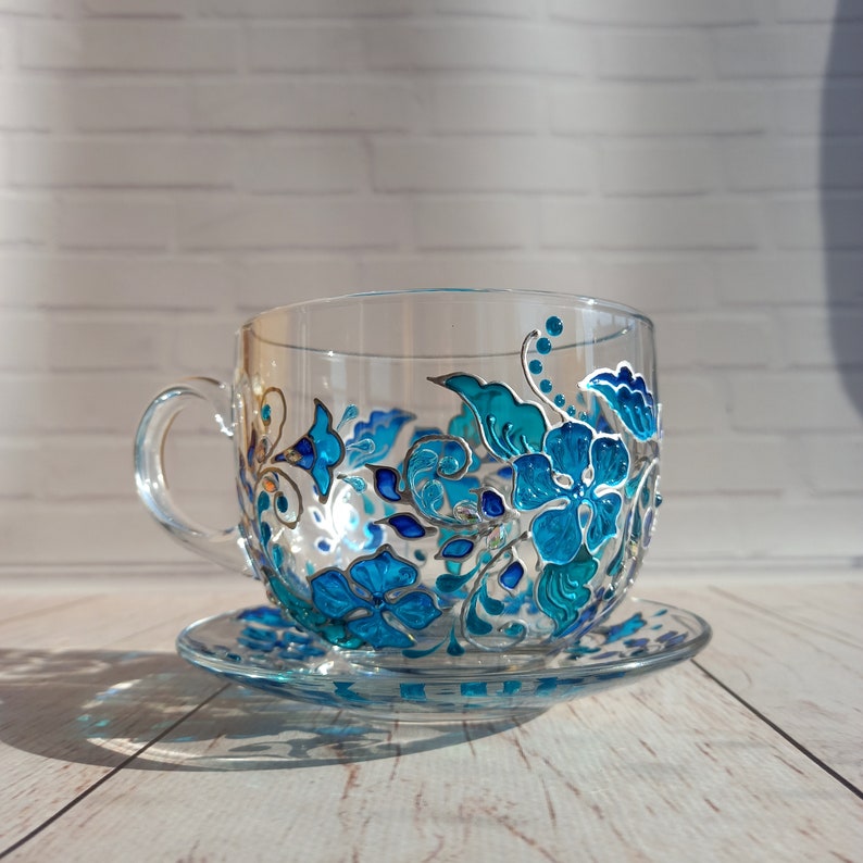 May include: A clear glass teacup and saucer set, hand-painted with a floral design in shades of turquoise and blue. The flowers and leaves are outlined in silver, creating a decorative and elegant look. The cup has a handle.