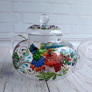 Puede incluir: Una tetera de vidrio transparente con tapa, con dise&ntilde;os florales y colibr&iacute;es pintados a mano en colores vibrantes. La tetera tiene un pico y un asa curvos, perfectos para servir t&eacute;. El dise&ntilde;o incluye tonos rojos, azules, verdes y amarillos.