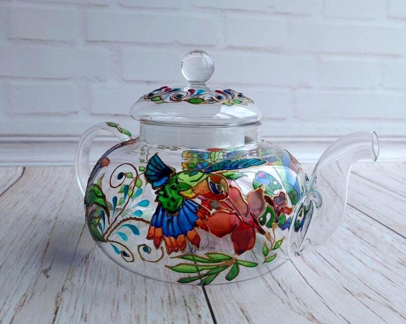 Puede incluir: Una tetera de vidrio transparente con tapa, con dise&ntilde;os florales y colibr&iacute;es pintados a mano en colores vibrantes. La tetera tiene un pico y un asa curvos, perfectos para servir t&eacute;. El dise&ntilde;o incluye tonos rojos, azules, verdes y amarillos.