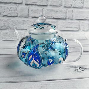 May include: A clear glass teapot with a lid and handle. The teapot is decorated with blue and white paisley and floral designs. The handle and spout are clear glass. The teapot sits on a white wooden surface.