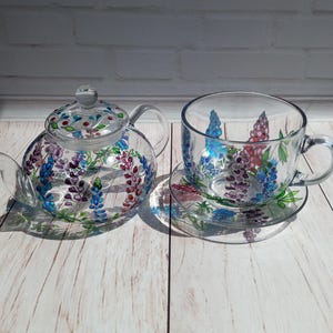 Puede incluir: Una tetera de cristal transparente con tapa a juego y una taza con platillo. El juego está decorado con un diseño floral en tonos azules, morados y rojos. La tetera tiene asa y boquilla.
